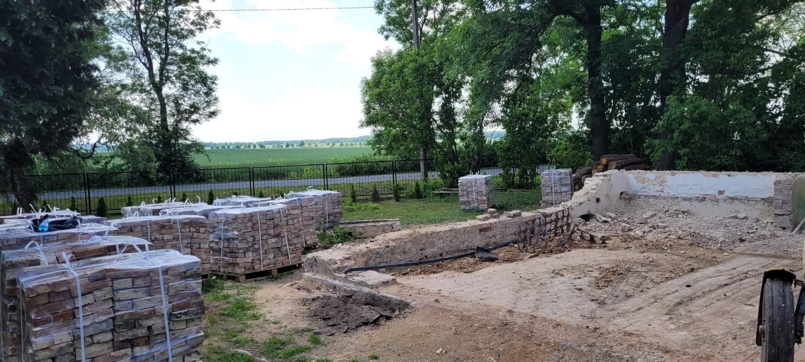 Częściowo rozebrana murowana konstrukcja na zewnątrz z paletami zapakowanych cegieł w tle, widok na zielone pole za ogrodzeniem.