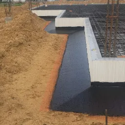 Zabezpieczone hydroizolacją fundamenty w trakcie budowy, widoczna warstwa izolacyjna na zewnętrznej ścianie fundamentu i zbrojenie płyty fundamentowej, obok wykop z piaskiem.