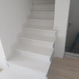 Białe, nowoczesne schody wewnętrzne z widocznymi fragmentami osoby montującej na górze, ukazujące etap wykończenia przy drzwiach.