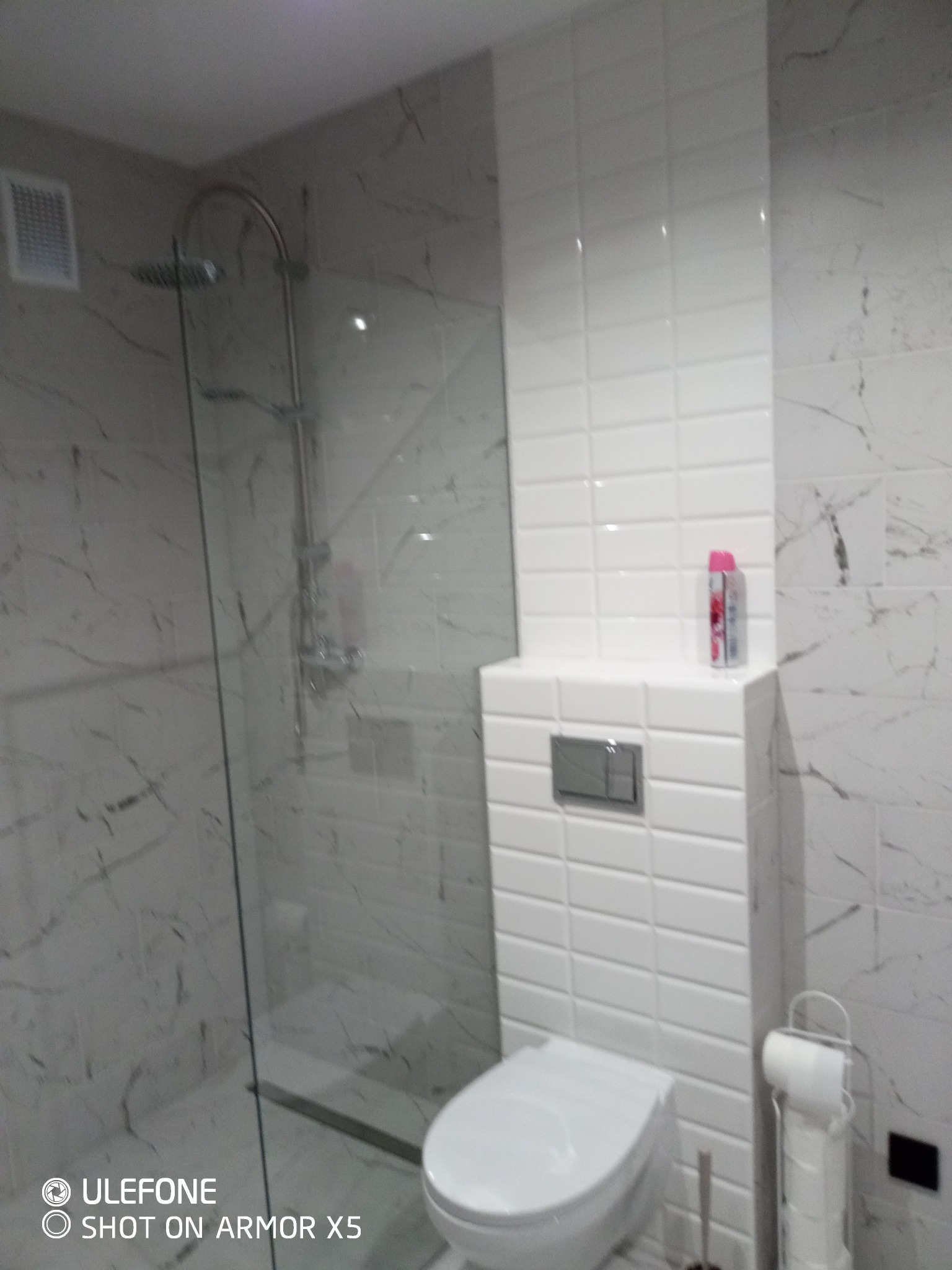 Nowoczesna łazienka z kabiną prysznicową typu walk-in, wykończona płytkami imitującymi marmur i białymi płytkami w kształcie cegiełek, z widoczną toaletą i akcesoriami.