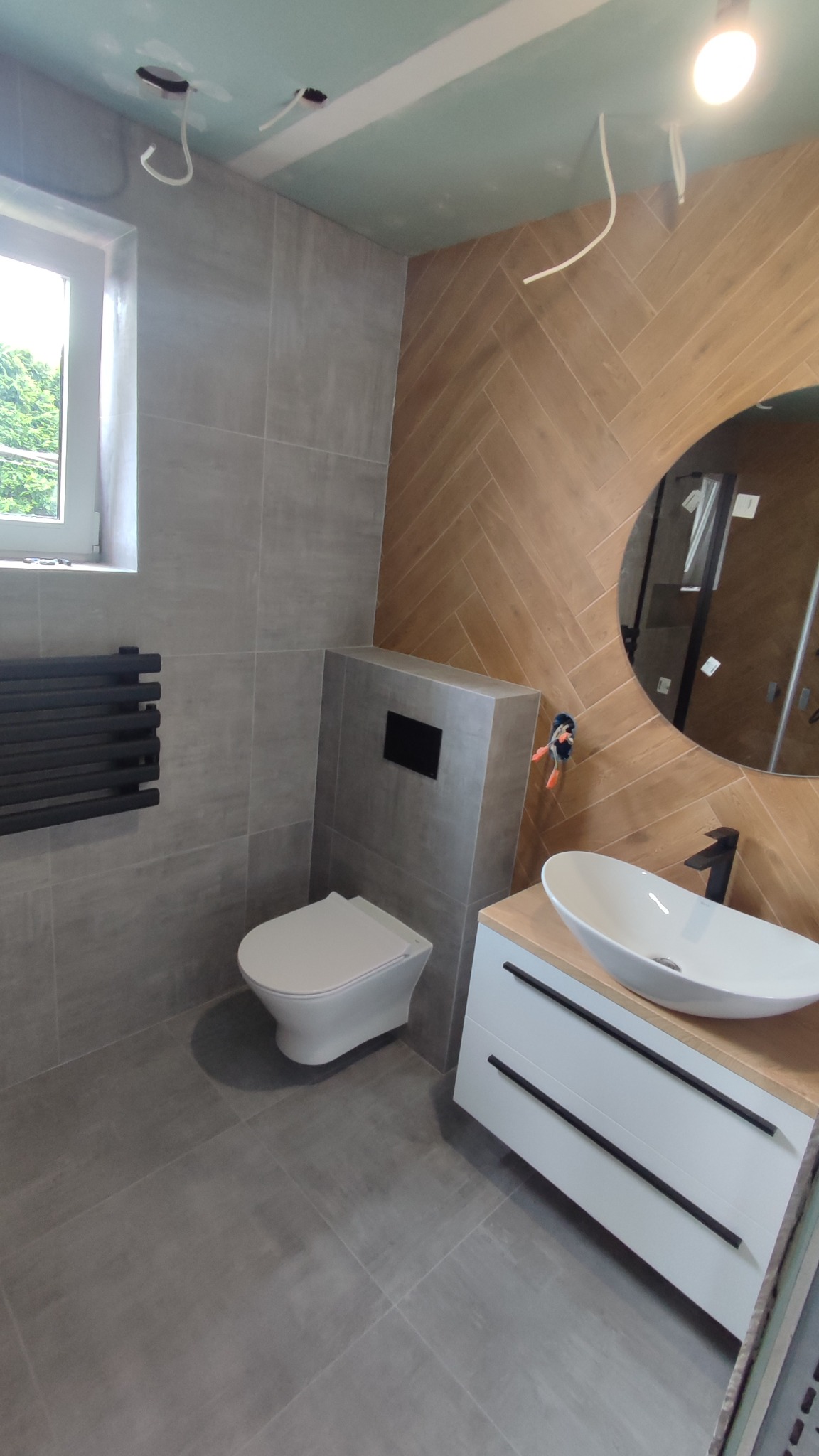 Nowoczesna łazienka z szarymi płytkami, drewnopodobnymi panelami ułożonymi w jodełkę, okrągłym lustrem, białą umywalką nablatową i wiszącą toaletą.