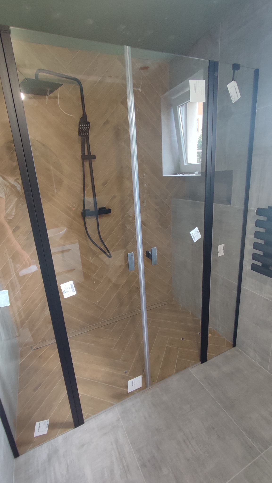 Kabina prysznicowa z czarnymi profilami, wykończona płytkami w jodełkę na ścianie prysznica i szarymi płytkami podłogowymi, z widoczną baterią prysznicową w kolorze czarnym i grzejnikiem drabinkowym.