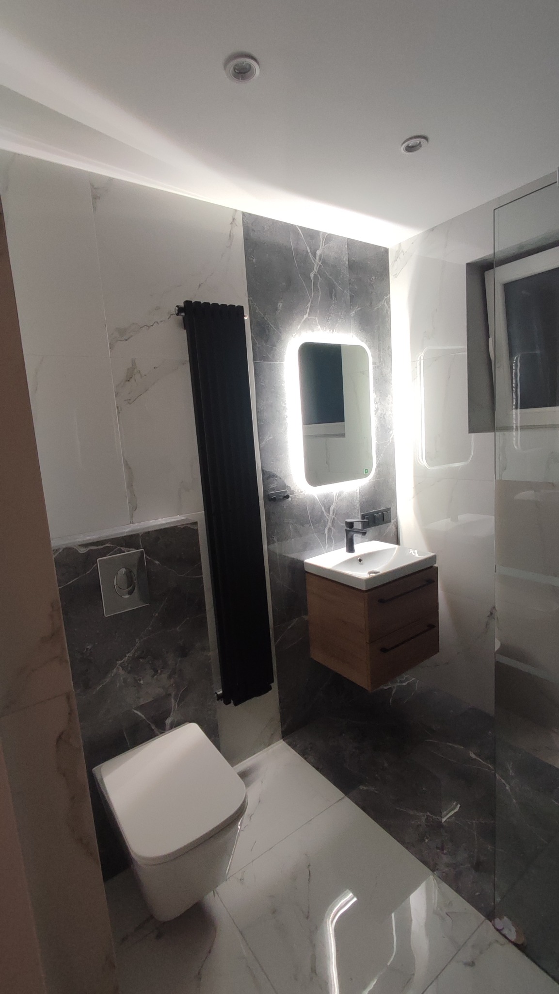 Nowoczesna łazienka z marmurowymi płytkami, podświetlanym lustrem i wiszącą szafką z umywalką, czarny grzejnik drabinkowy i wisząca toaleta.