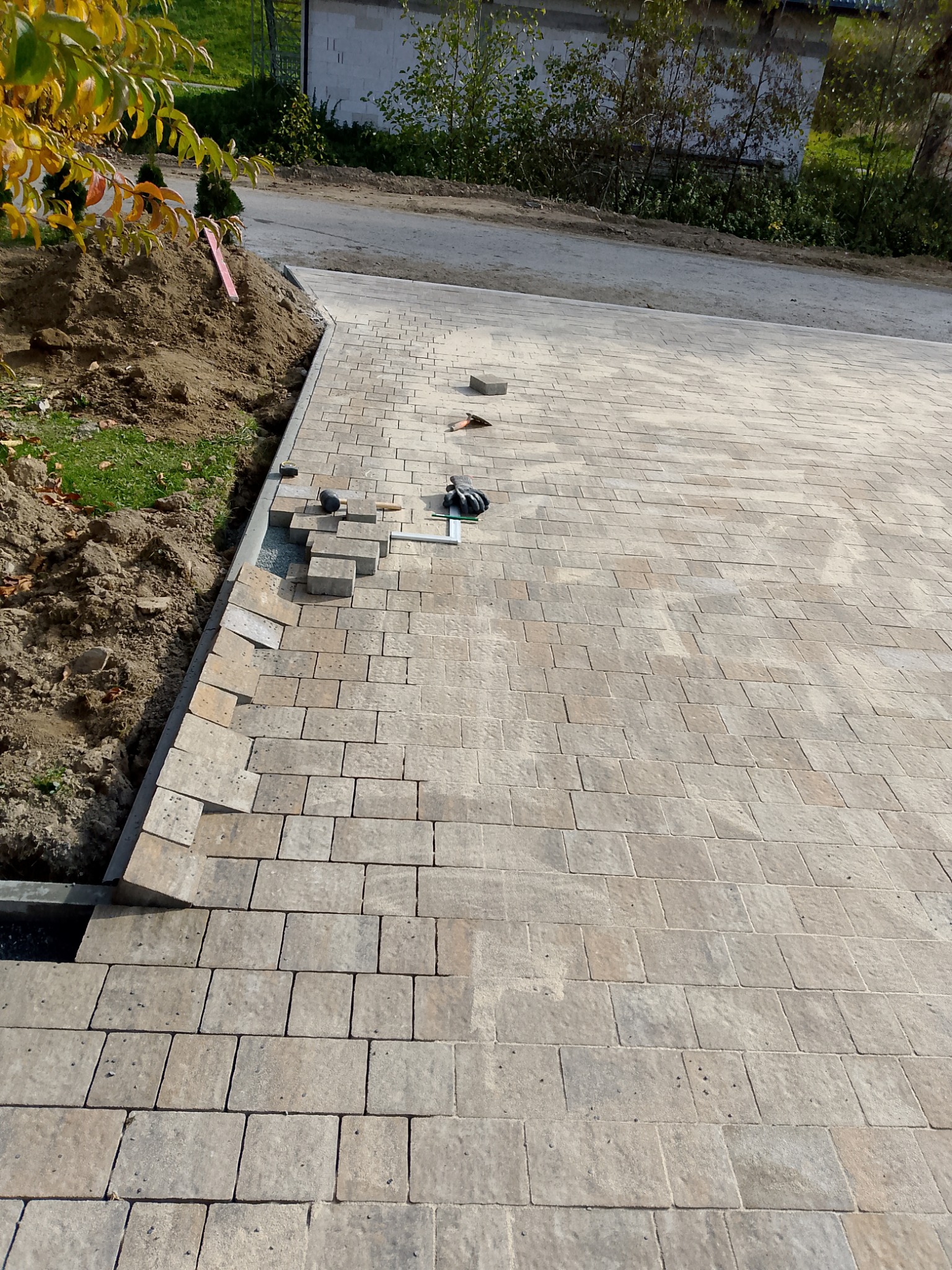Układanie beżowej kostki brukowej na podjeździe, widoczne narzędzia i fragmenty niedokończonej nawierzchni przy krawędzi. W tle zieleń i budynek.