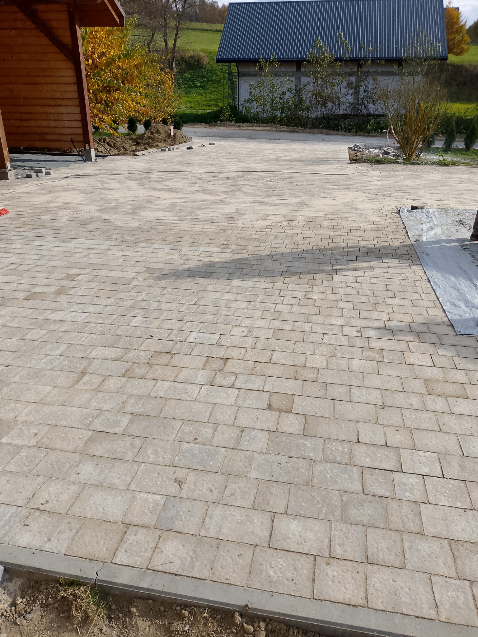Jasnoszara kostka brukowa ułożona na podjeździe, z widocznymi elementami krajobrazu i fragmentem drewnianej konstrukcji po lewej stronie. Częściowo zakończone prace.