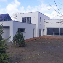 Nowoczesny, biały dom z płaskim dachem i drewnianym tarasem, widok z ogrodu z młodymi iglakami i żółtym wężem ogrodowym na ziemi.