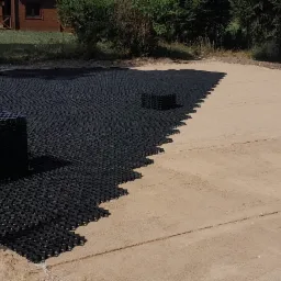 Częściowo ułożona, czarna, plastikowa geokrata na przygotowanym, piaszczystym podłożu, z widoczną stertą nieużytych elementów, teren przygotowany pod utwardzenie drogi.