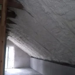 Poddasze ocieplone szarą pianką poliuretanową, widoczna drewniana konstrukcja więźby dachowej i okno w szczycie ściany.