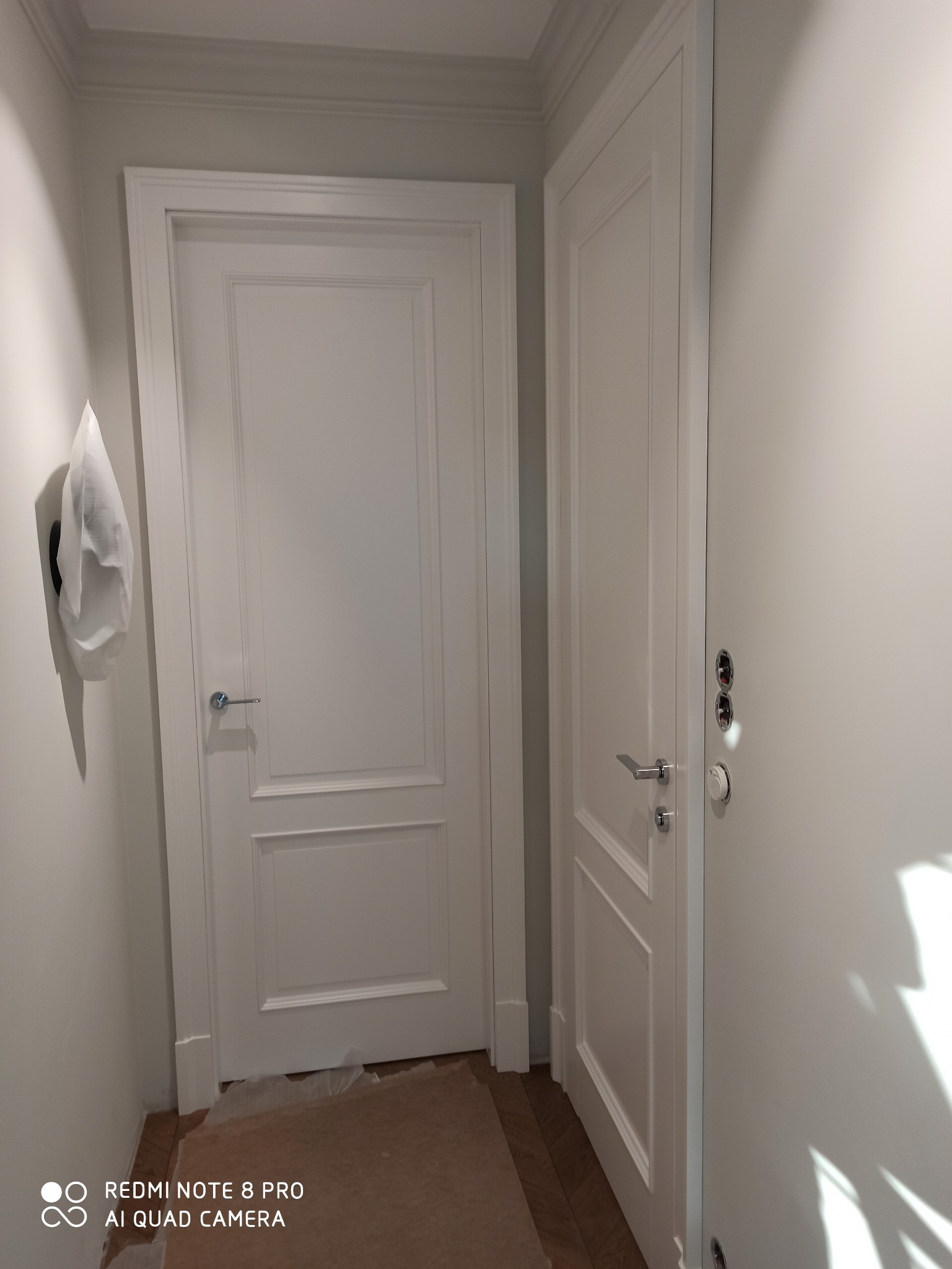 Dwa białe, panelowe drzwi wewnętrzne z chromowanymi klamkami w jasnym korytarzu, podłoga zabezpieczona kartonem.