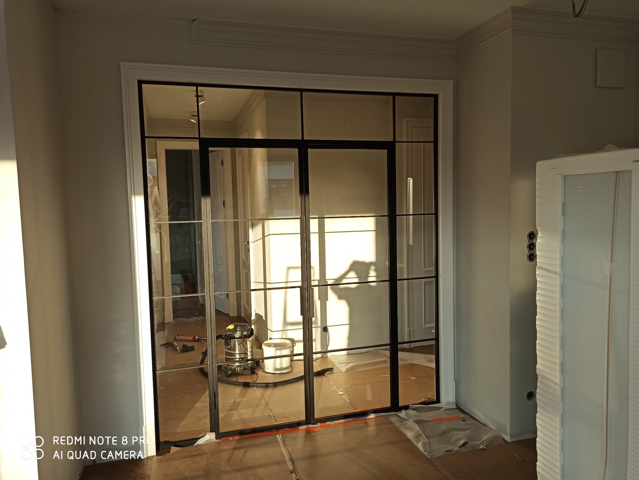 Montaż szklanych drzwi loftowych z czarnymi szprosami, widoczny odkurzacz i narzędzia na podłodze zabezpieczonej kartonem, cień osoby na szybie.