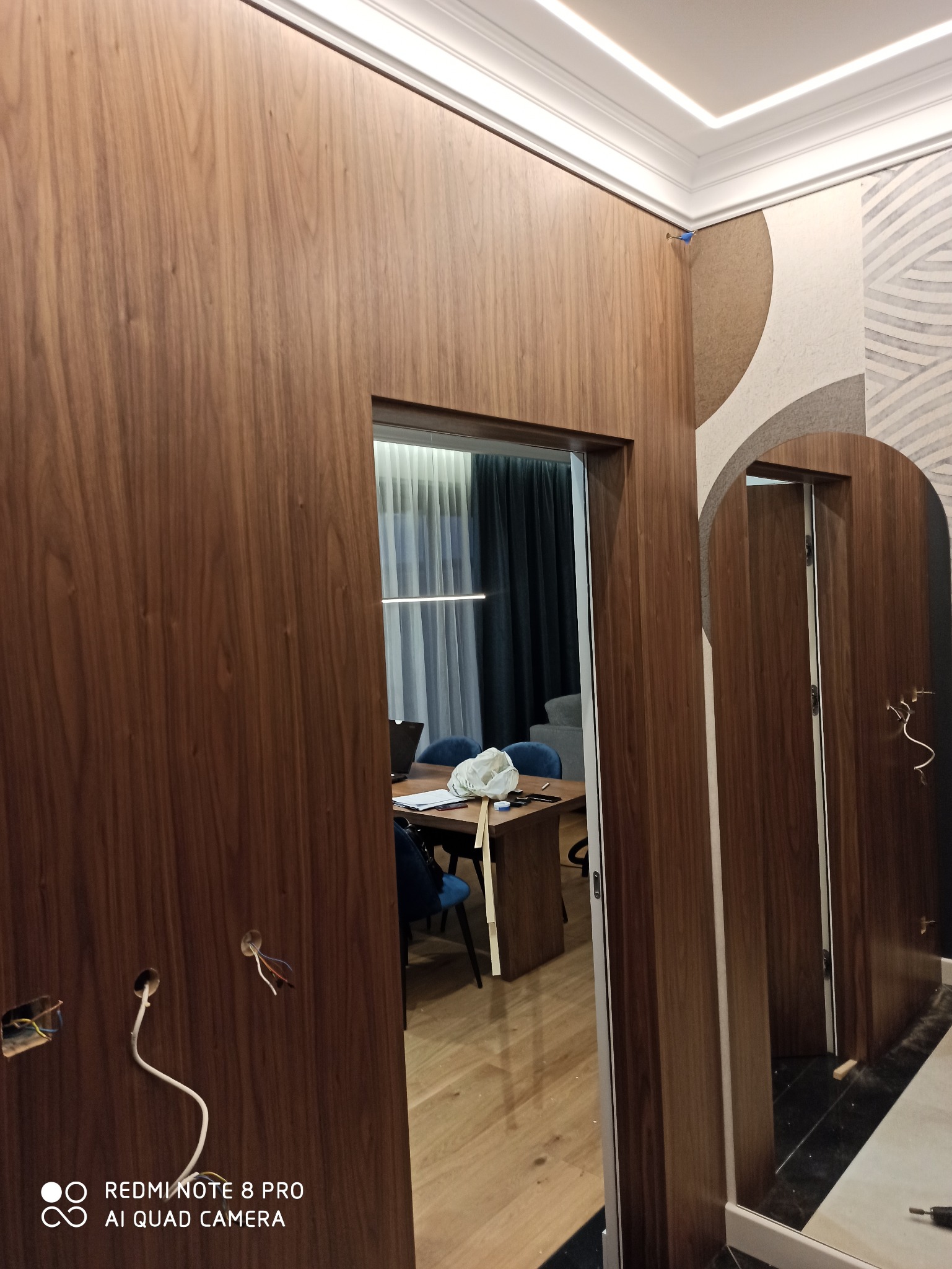 Wykończenie wnętrza: drewniana boazeria ścienna z ukrytymi drzwiami, widoczne otwory na instalacje elektryczne i fragment wnętrza pokoju biurowego z biurkiem i krzesłami w tle.