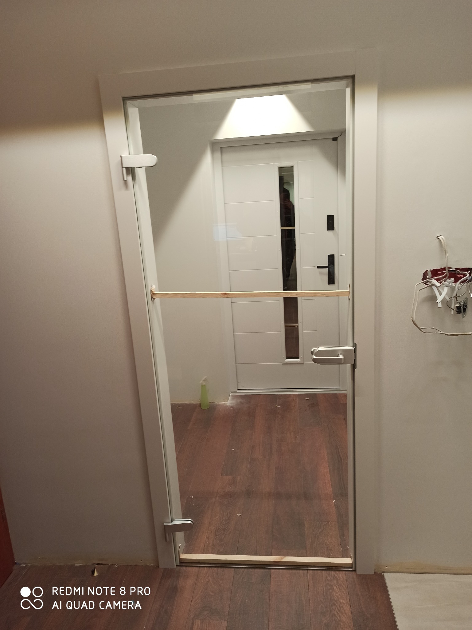 Szklane drzwi wewnętrzne w trakcie montażu, zabezpieczone drewnianą listwą, widoczna instalacja elektryczna na ścianie.