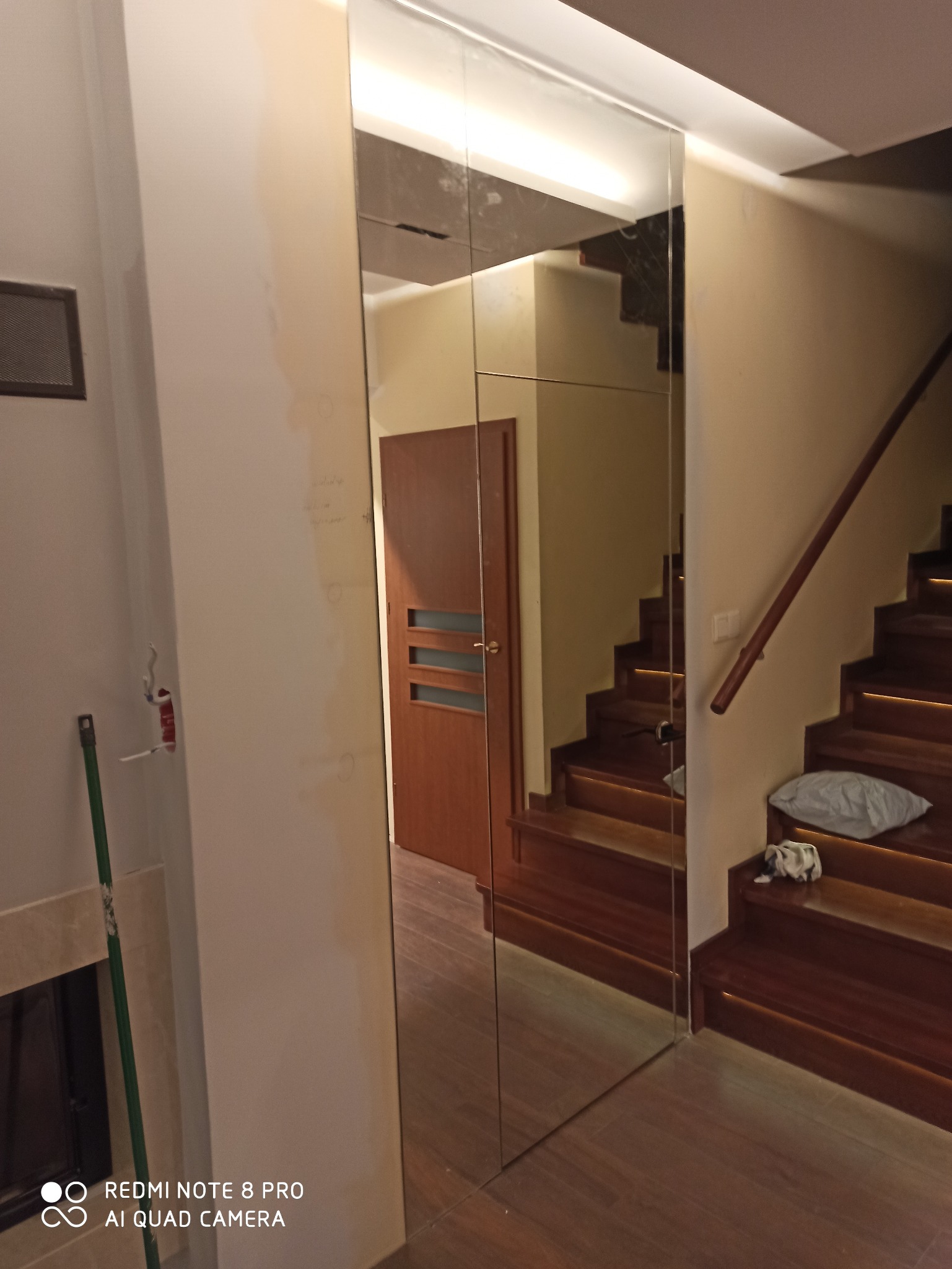 Wnętrze mieszkania z zamontowanymi lustrzanymi drzwiami przesuwnymi, odbijającymi drewniane schody z podświetleniem LED i brązowe drzwi z trzema szybkami.