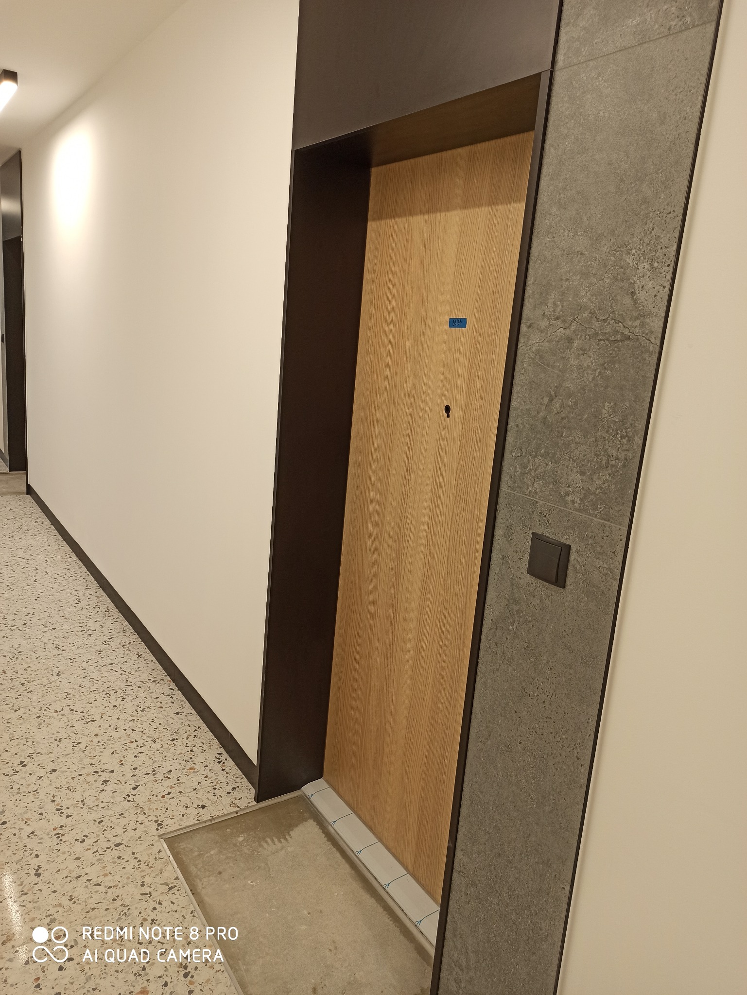 Nowoczesne drzwi wejściowe z drewnianą okleiną i szarą ościeżnicą, widoczne w korytarzu z podłogą lastryko, z małą niebieską naklejką na drzwiach i czarnym włącznikiem światła na ścianie obok.