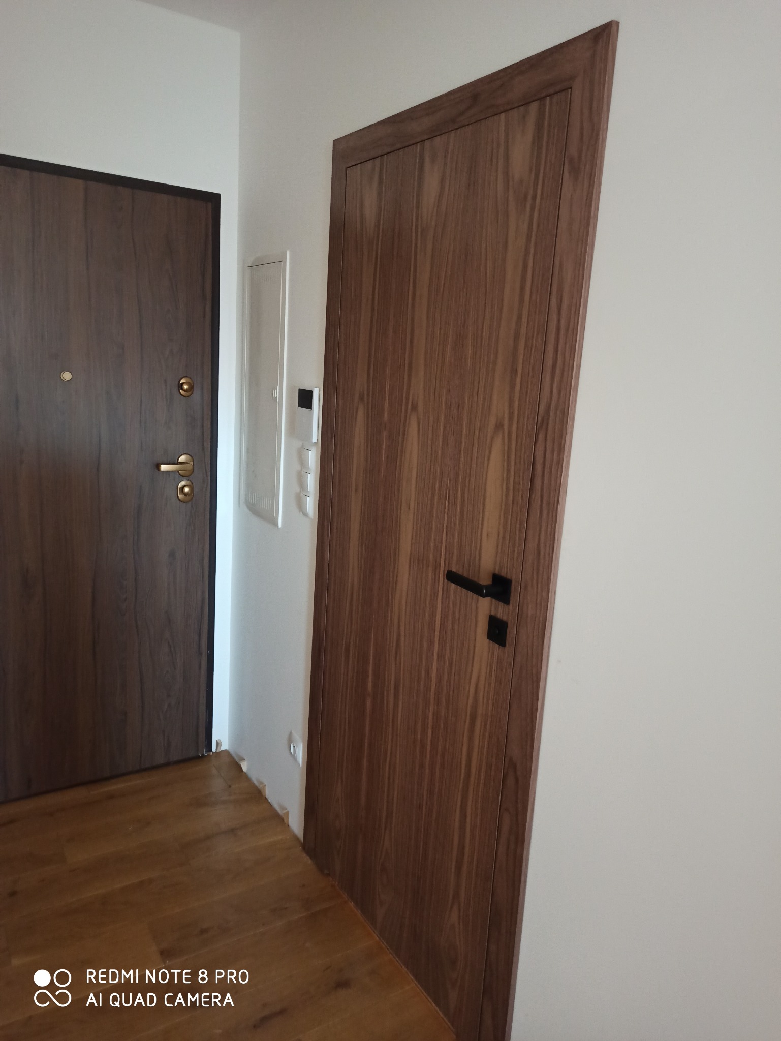 Dwa rodzaje drzwi w przedpokoju: brązowe drzwi wejściowe z okuciami w kolorze złota oraz wewnętrzne drzwi z okleiną drewnopodobną i czarną klamką.