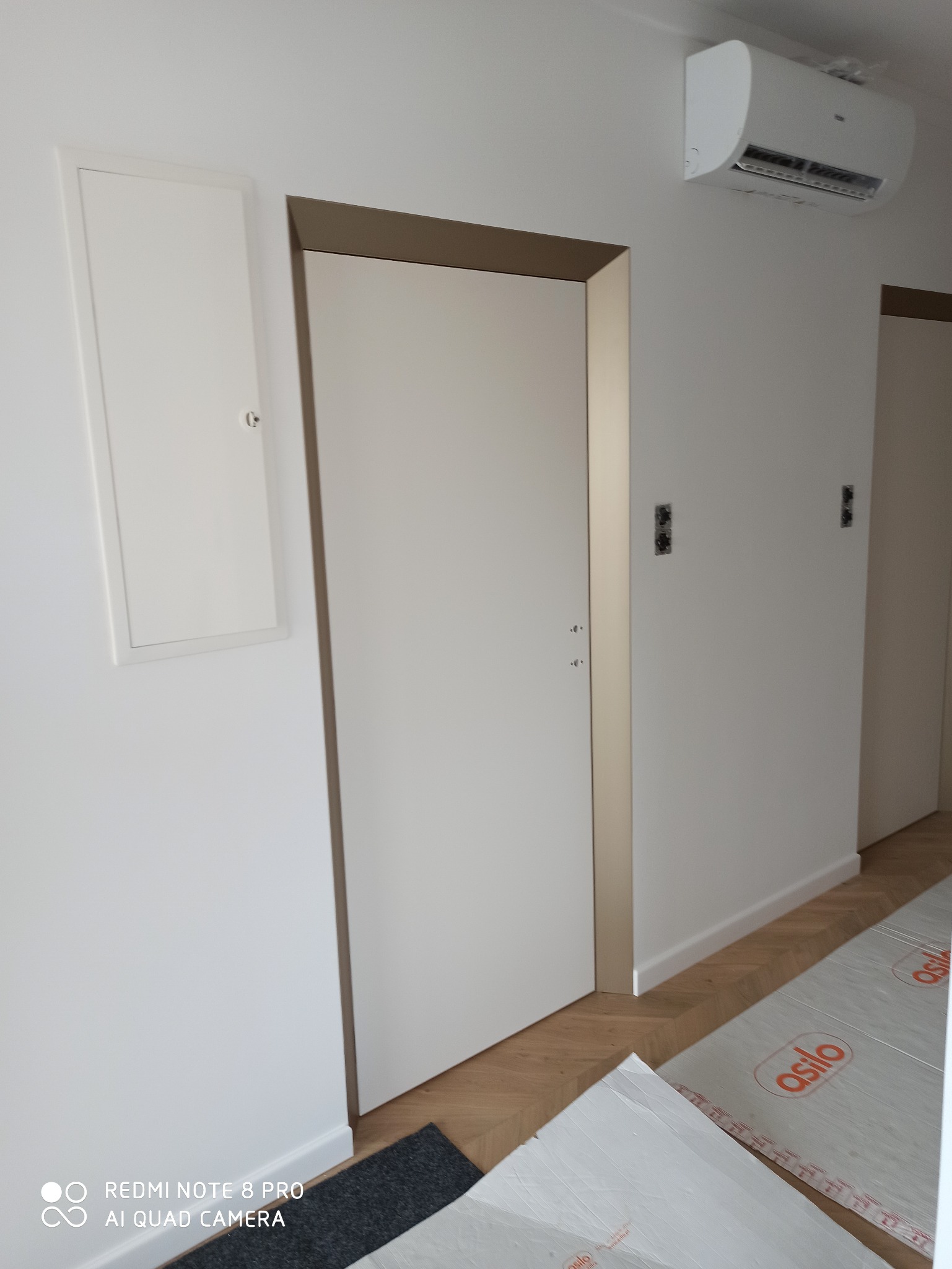 Białe, minimalistyczne drzwi wewnętrzne z brązową ramą, zamontowane w białym pomieszczeniu z parkietem i klimatyzatorem na ścianie. Podłoga zabezpieczona folią ochronną.