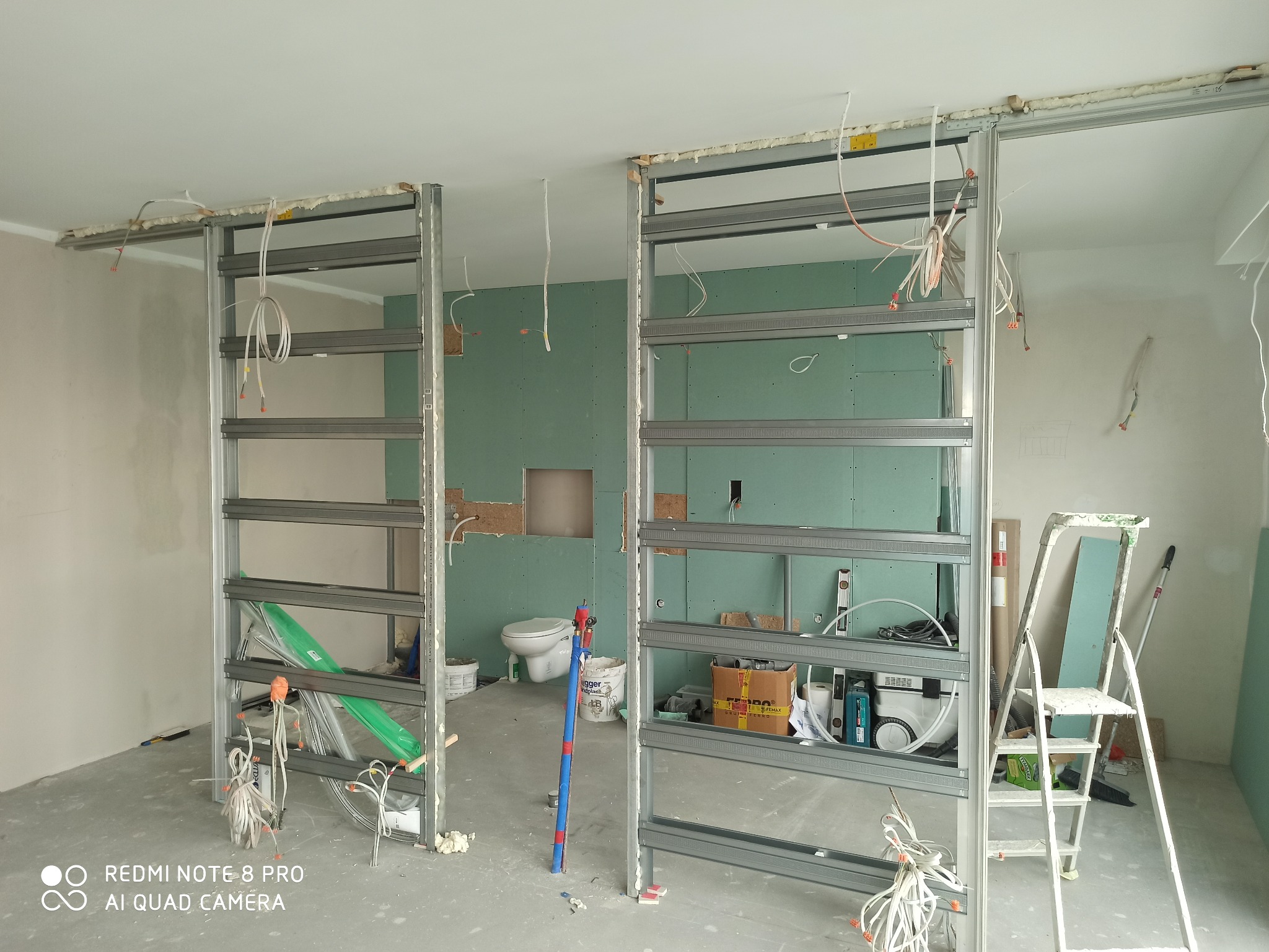 Montaż drzwi przesuwnych: widoczne metalowe profile konstrukcyjne z zamontowanymi szynami sufitowymi, instalacja elektryczna w trakcie prowadzenia, ściany z płyt gipsowo-kartonowych, w tle toaleta...