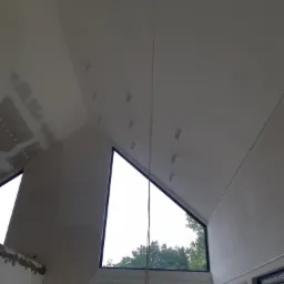 Wnętrze w Olkuszu w trakcie wykańczania: widoczne ściany po gładziowaniu i przygotowaniu do malowania, zamontowane punkty oświetleniowe na skośnym suficie oraz duże okna z widokiem na zieleń.