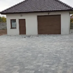 Szeroki podjazd z szarej kostki brukowej prowadzący do garażu z brązową bramą, obok betonowy płot i rząd tuj.