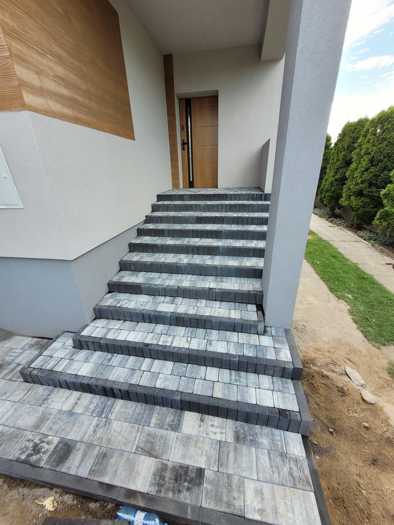 Schody wejściowe do domu wyłożone szaro-białą kostką brukową, z ciemnoszarym obramowaniem, prowadzące do drewnianych drzwi.