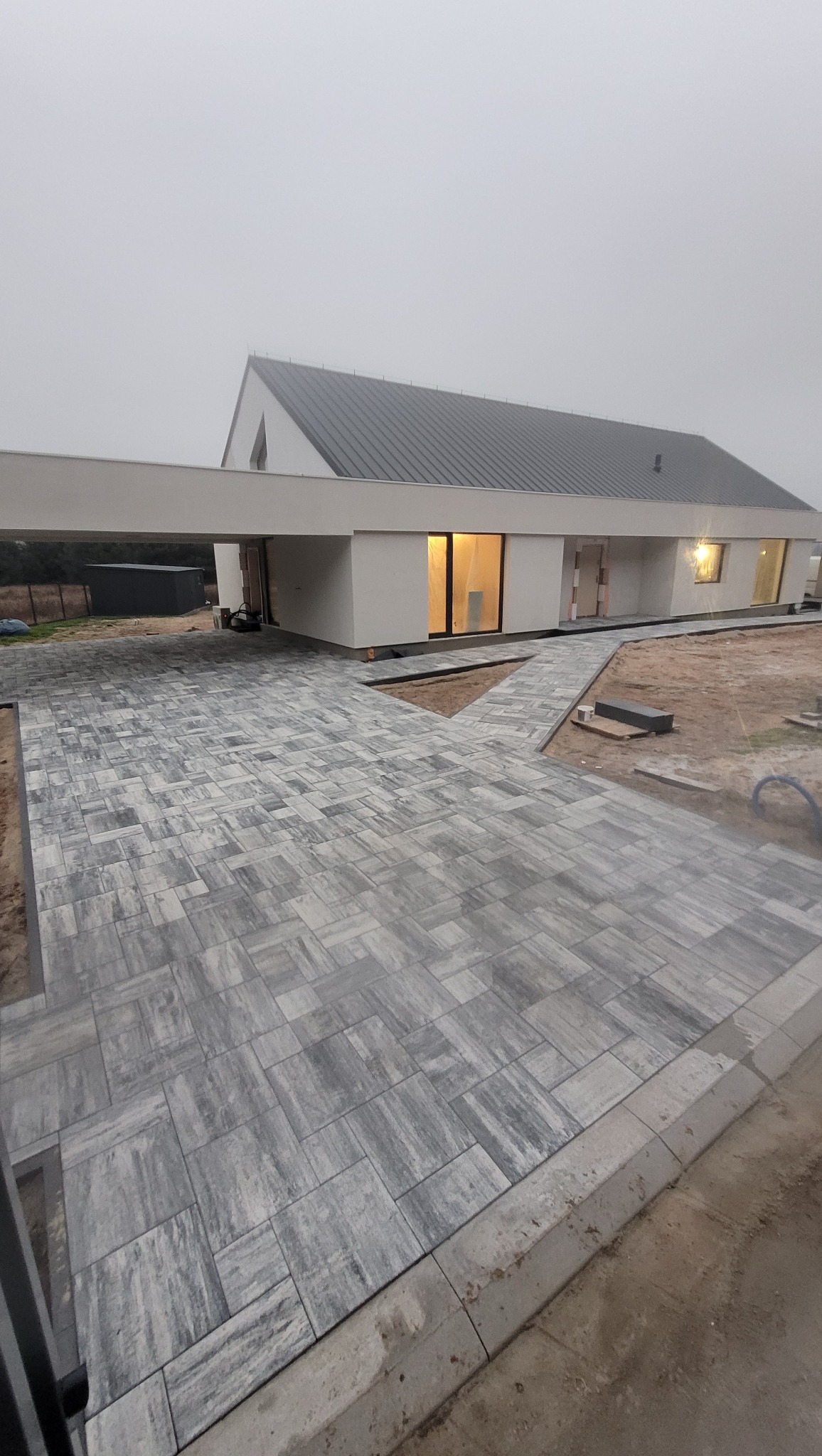 Ułożona szara kostka brukowa prowadząca do nowoczesnego domu z płaskim dachem w pochmurny dzień. Widoczny fragment chodnika z kostki i podjazd.