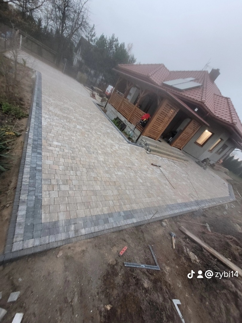 Ukończony podjazd z kostki brukowej w odcieniach beżu i szarości prowadzący do domu z drewnianą werandą, widoczne narzędzia brukarskie na ziemi.