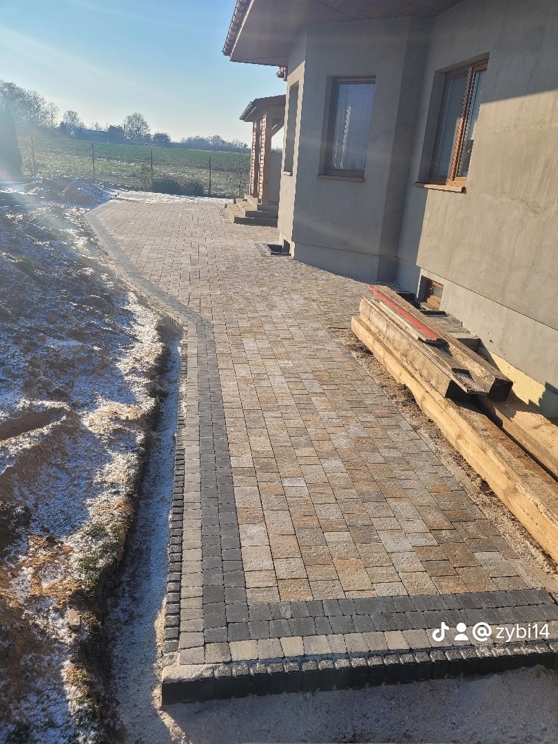 Ułożona ścieżka z kostki brukowej w odcieniach beżu i szarości prowadząca wzdłuż ściany domu, z widocznymi deskami opartymi o ścianę i fragmentem ogrodu w tle.