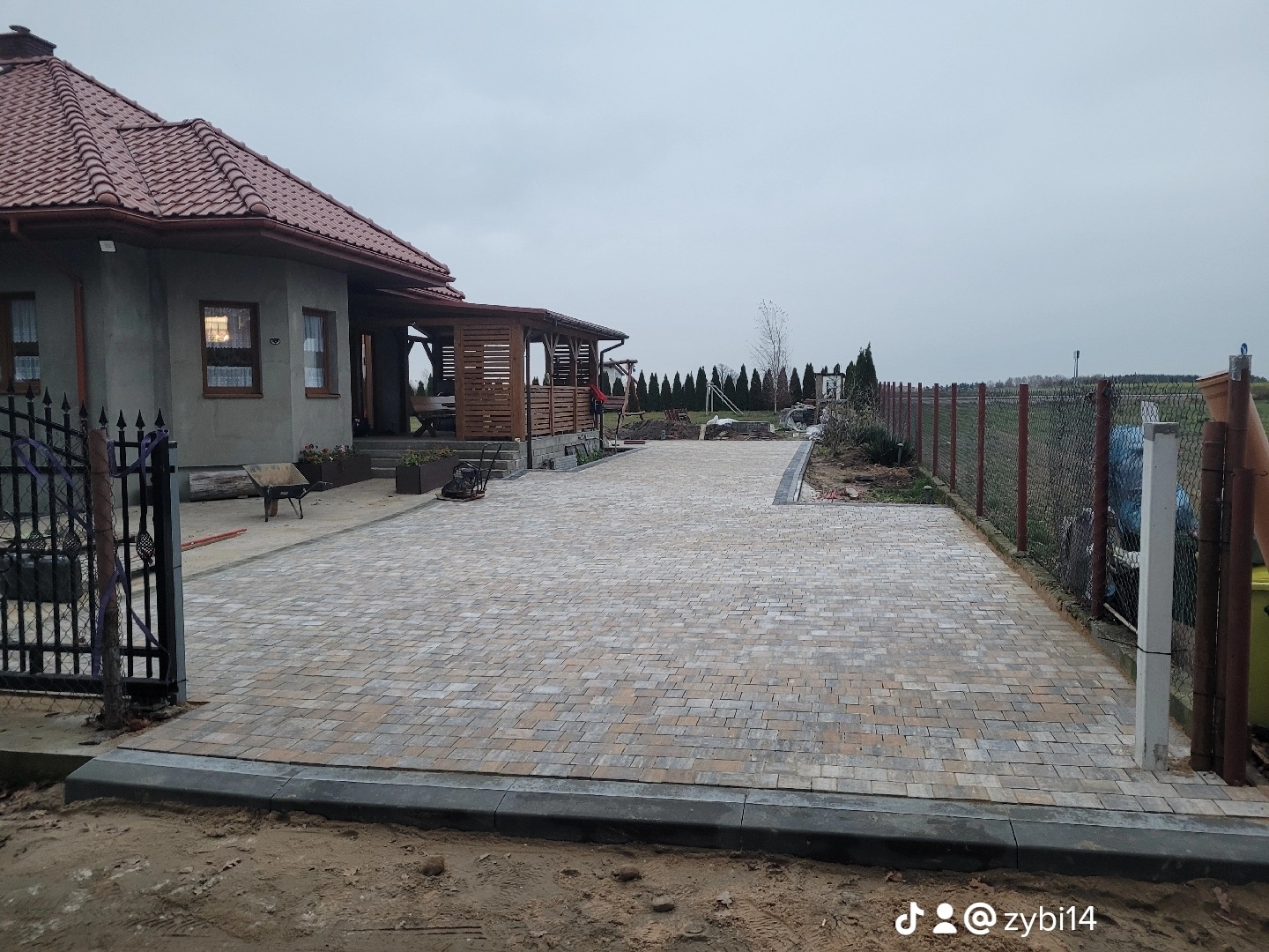 Szeroki podjazd z kostki brukowej w odcieniach beżu i szarości prowadzący do domu z drewnianą altaną, widok zza czarnej, metalowej bramy wjazdowej, w tle rząd tuj.