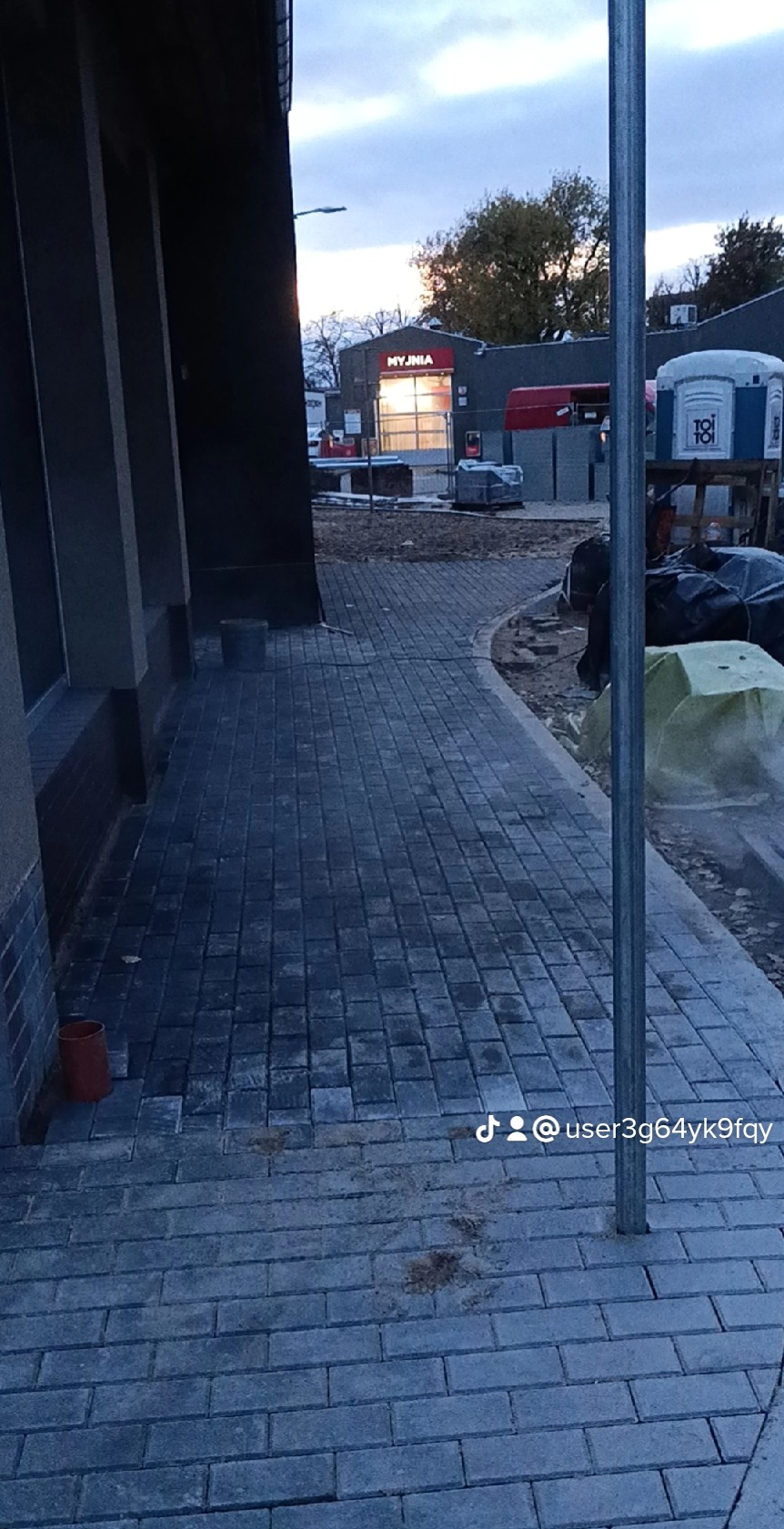 Ułożona szara kostka brukowa tworząca chodnik, widoczny budynek myjni w tle i tymczasowa toaleta przenośna.