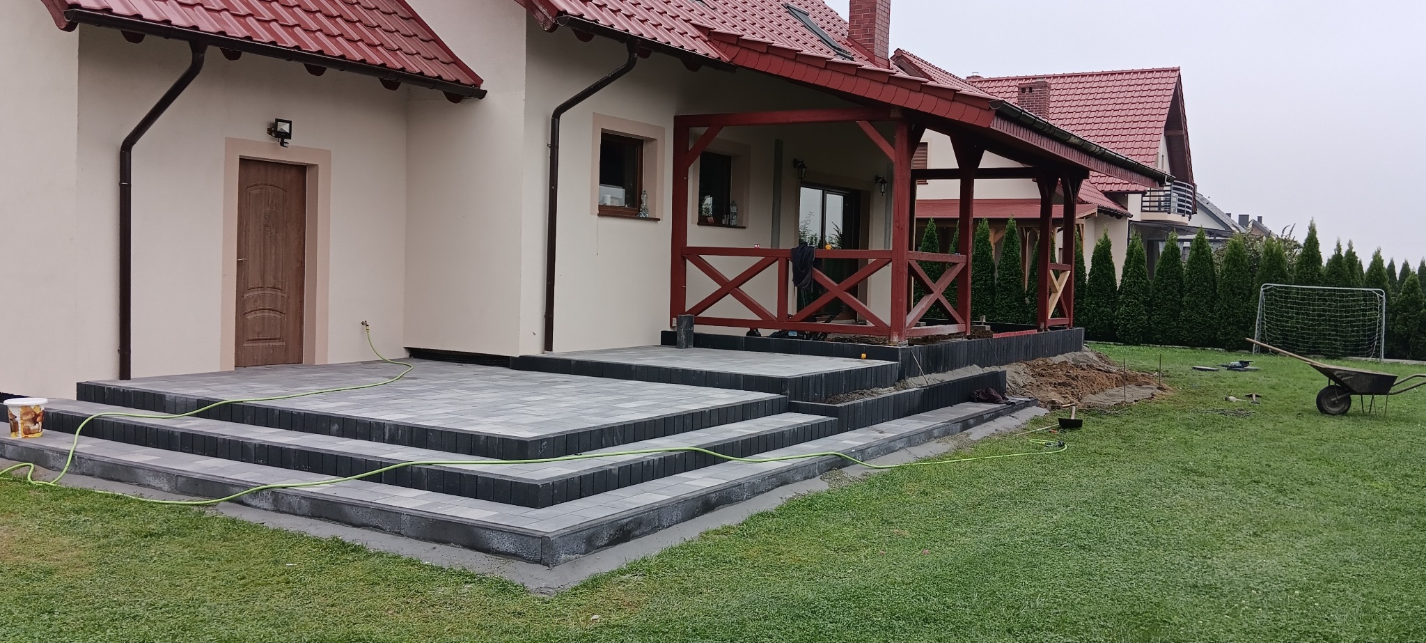 Schody wejściowe do domu wyłożone szarą kostką brukową, widoczny fragment trawnika, wąż ogrodowy i taczka z narzędziami.