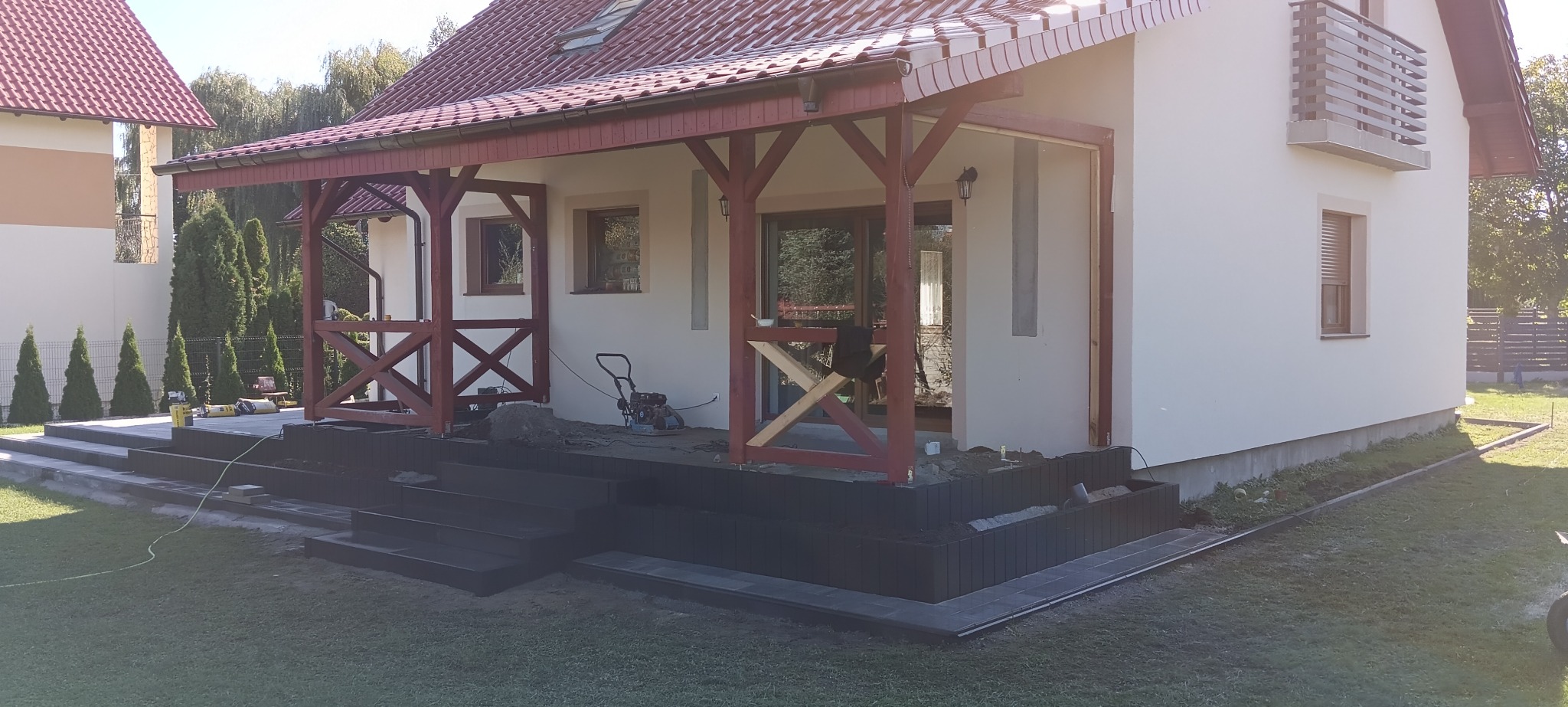 Montaż stopni z ciemnej kostki brukowej przed wejściem do domu z drewnianą werandą, widoczna zagęszczarka i materiały budowlane.