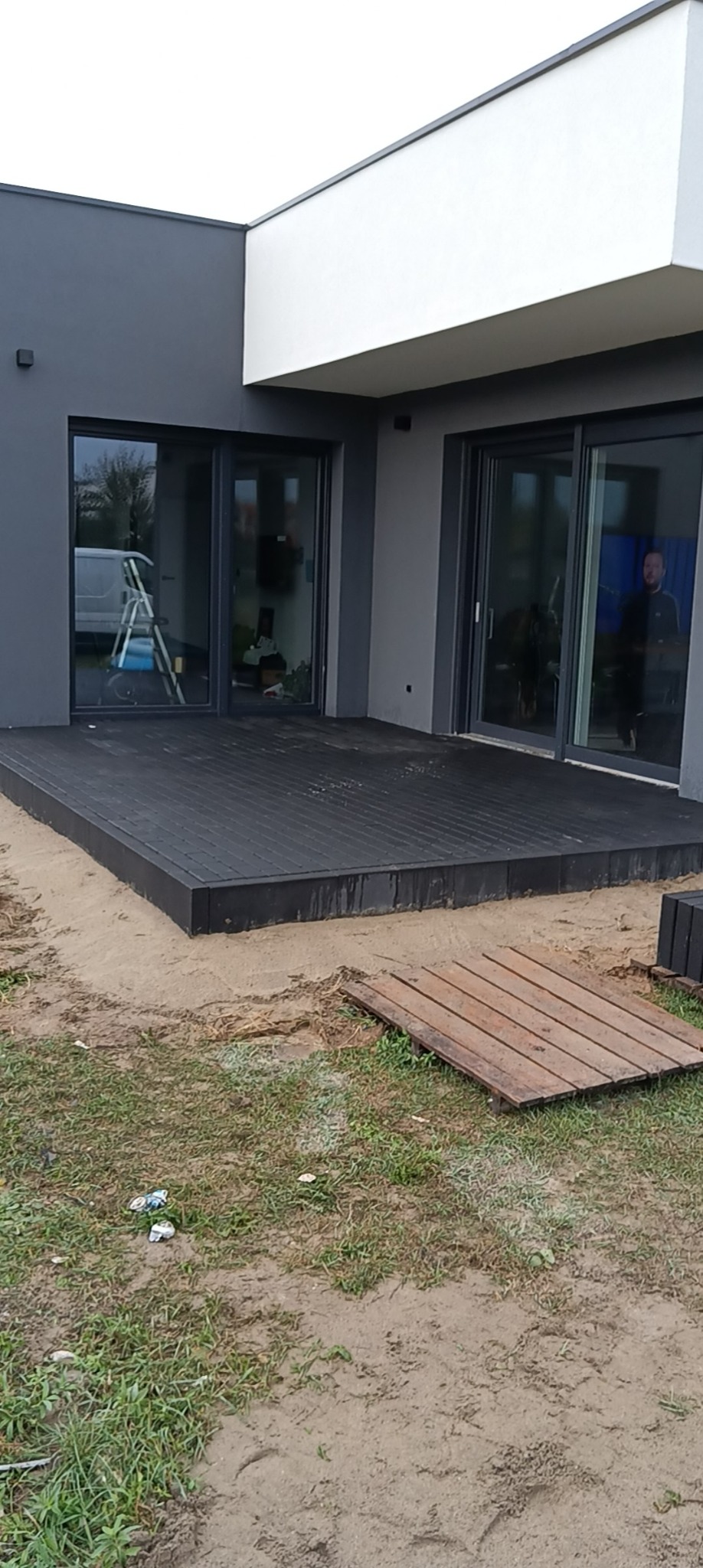 Nowoczesny taras z ciemnej kostki kompozytowej przylegający do budynku z przesuwnymi drzwiami, widoczny fragment trawnika i piasku z elementami porządkowymi.