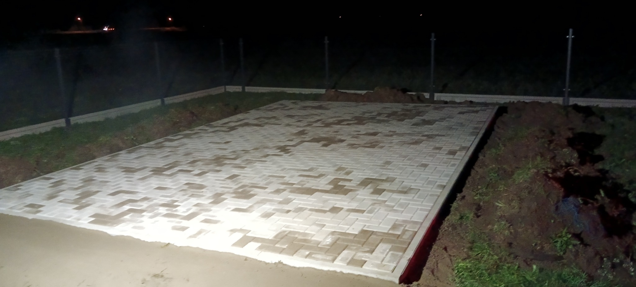 Ułożona jasnoszara kostka brukowa w jodełkę na przygotowanym podłożu, widoczne wykopane obrzeża i słupy ogrodzenia w tle, nocne oświetlenie.