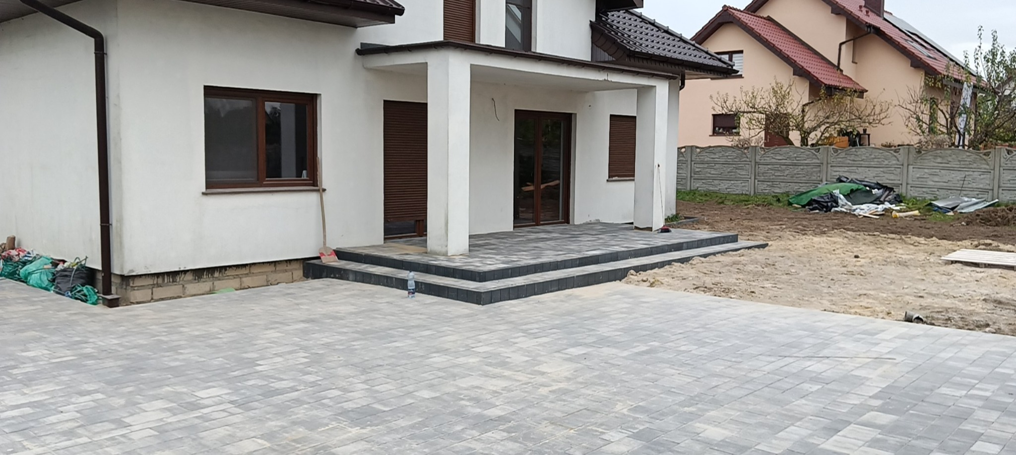 Ułożona szara kostka brukowa przed domem z białymi kolumnami i trzema stopniami wejściowymi, widoczny fragment ogrodu i sąsiedni dom.