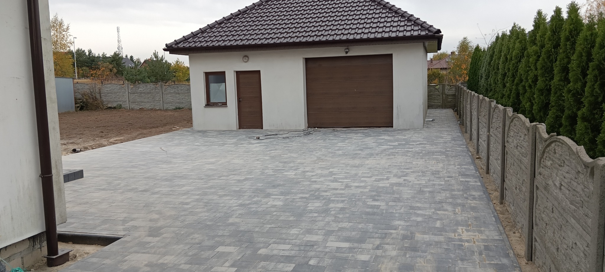 Szeroki podjazd z szarej kostki brukowej prowadzący do garażu z brązową bramą, obok betonowy płot i rząd tuj.