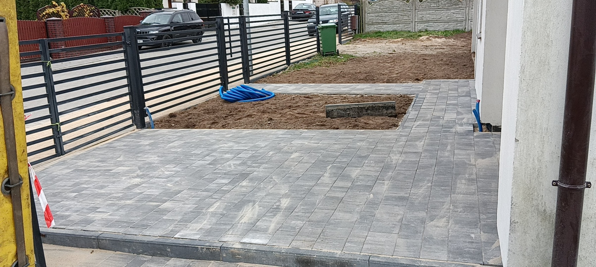 Ułożona szara kostka brukowa na podjeździe, z przygotowanymi fragmentami pod dalsze prace, widoczny zwój niebieskiego węża i metalowe ogrodzenie posesji.