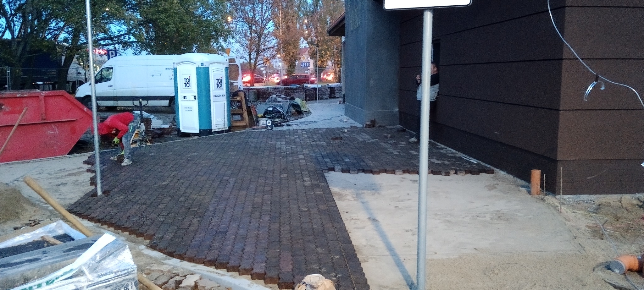 Układanie ciemnej kostki brukowej o falistym wzorze, widoczny pracownik w czerwonej bluzie, w tle biały van i przenośne toalety.