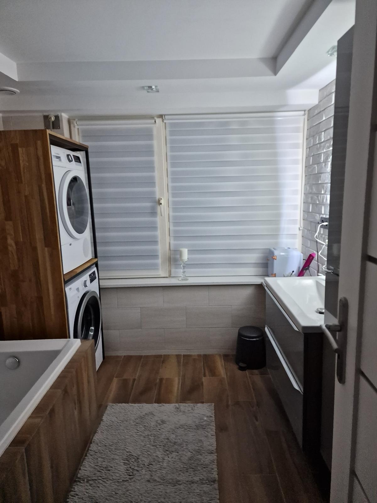 Ukończona łazienka z drewnianą podłogą, szarym dywanikiem, pralką i suszarką w drewnianej zabudowie, białą umywalką z szarą szafką oraz żaluzjami okiennymi.