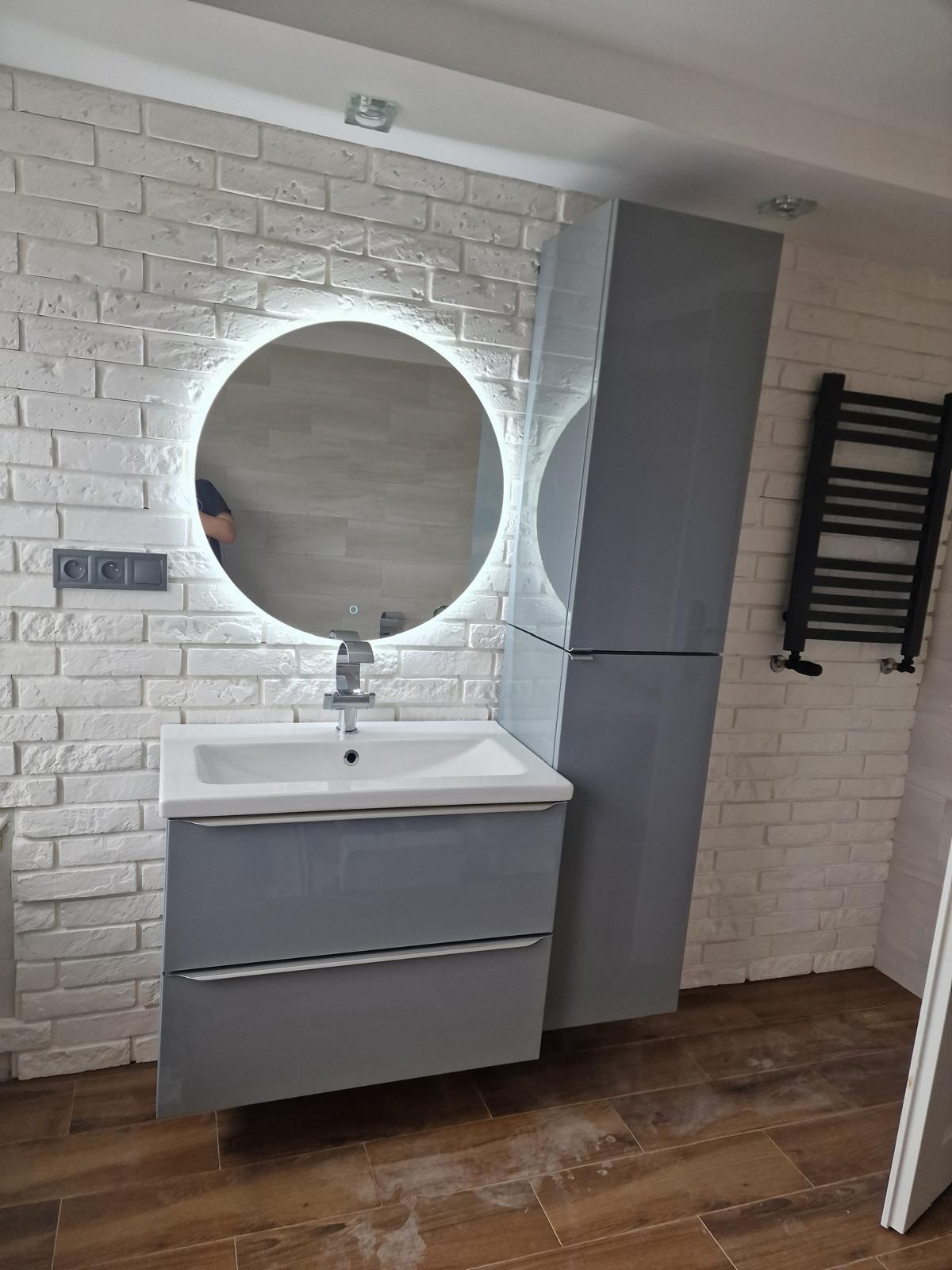 Nowoczesna łazienka z umywalką na szafce w kolorze szarym, okrągłym lustrem z podświetleniem LED, grzejnikiem drabinkowym i ścianą z białej cegły.