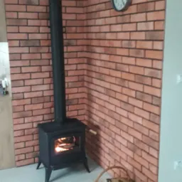 Czarne żeliwne palenisko z płomieniami widocznymi przez szybę, ustawione przy ścianie wyłożonej cegłą, z koszem drewna obok, a na ścianie zegar.