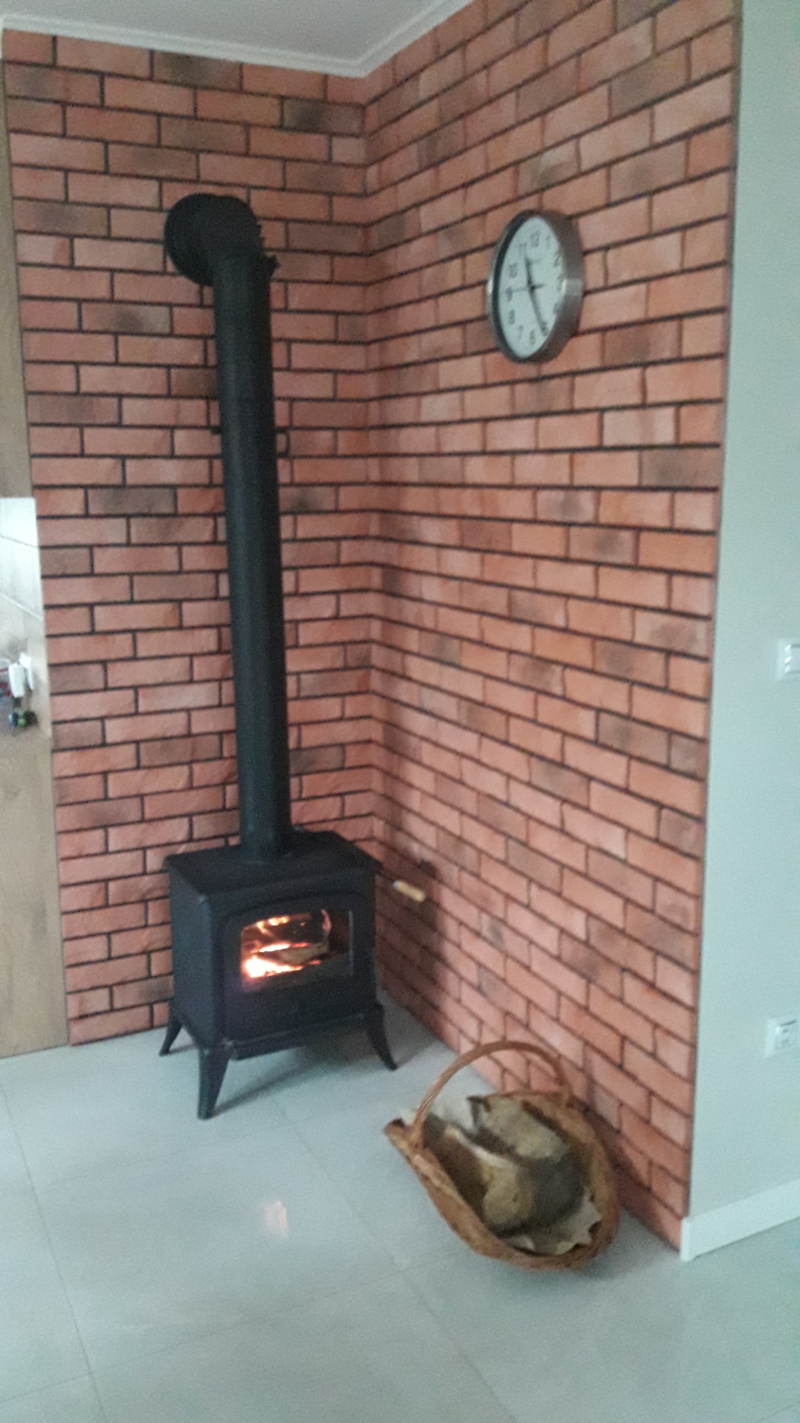 Czarne żeliwne palenisko z płomieniami widocznymi przez szybę, ustawione przy ścianie wyłożonej cegłą, z koszem drewna obok, a na ścianie zegar.