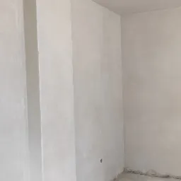 Surowe, jasne wnętrze po nałożeniu tynku, widoczny pionowy element konstrukcyjny oraz niedoskonałości na podłodze i ścianach.