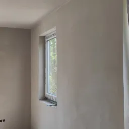 Wnętrze pomieszczenia po nałożeniu szarej warstwy wykończeniowej na ściany i sufit, widoczne okno z widokiem na zieleń, surowy beton na podłodze, ślady niedokończonych prac przy listwach...