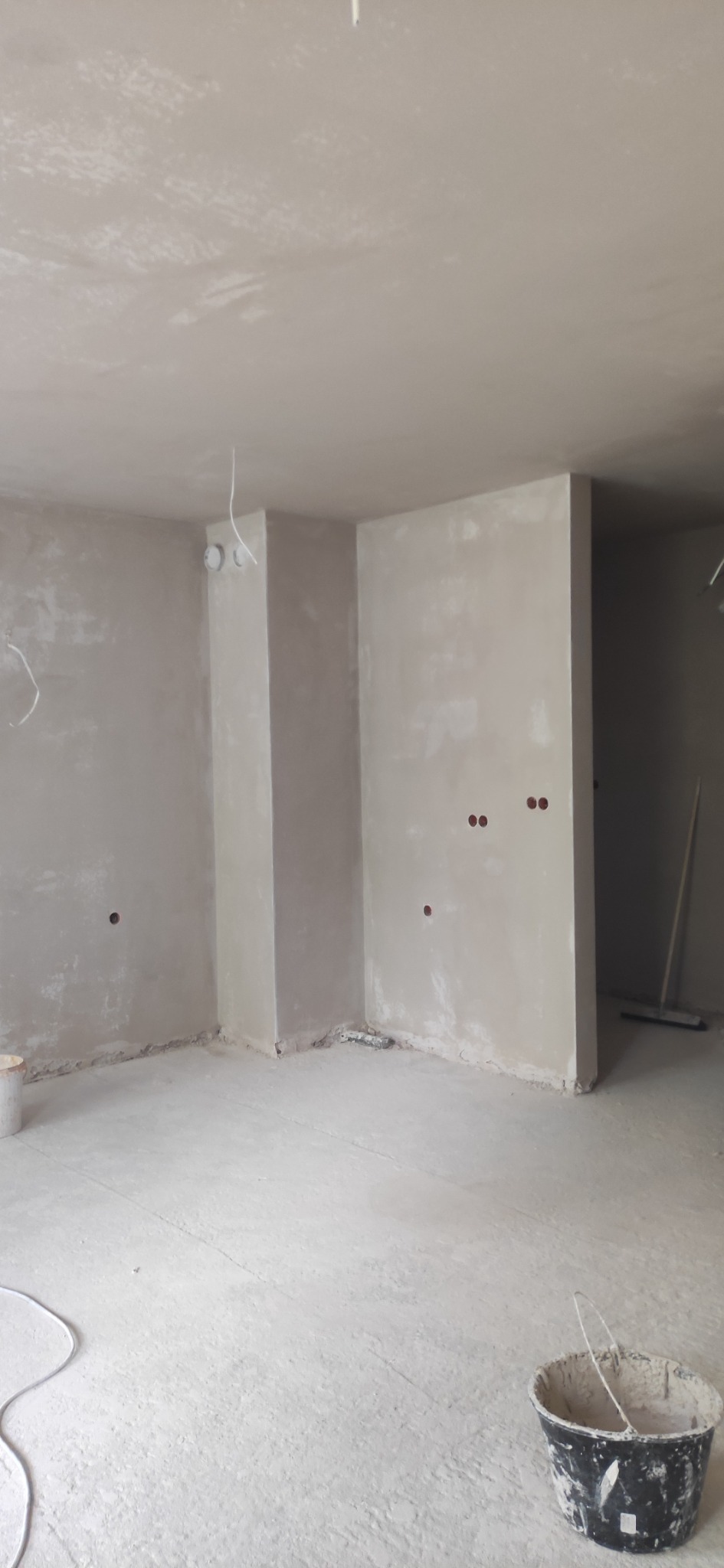 Surowe, szare wnętrze w trakcie prac wykończeniowych, ściany pokryte świeżo nałożoną warstwą masy, widoczne puszki instalacyjne i wiadro z zaprawą na podłodze.
