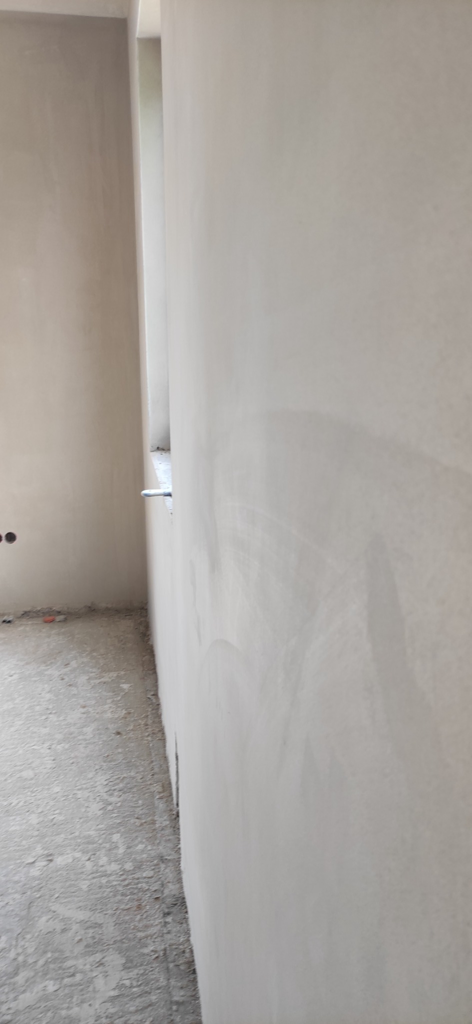 Świeżo położona, jasnoszara warstwa na ścianie w trakcie prac wykończeniowych, widoczny fragment okna i surowa podłoga.