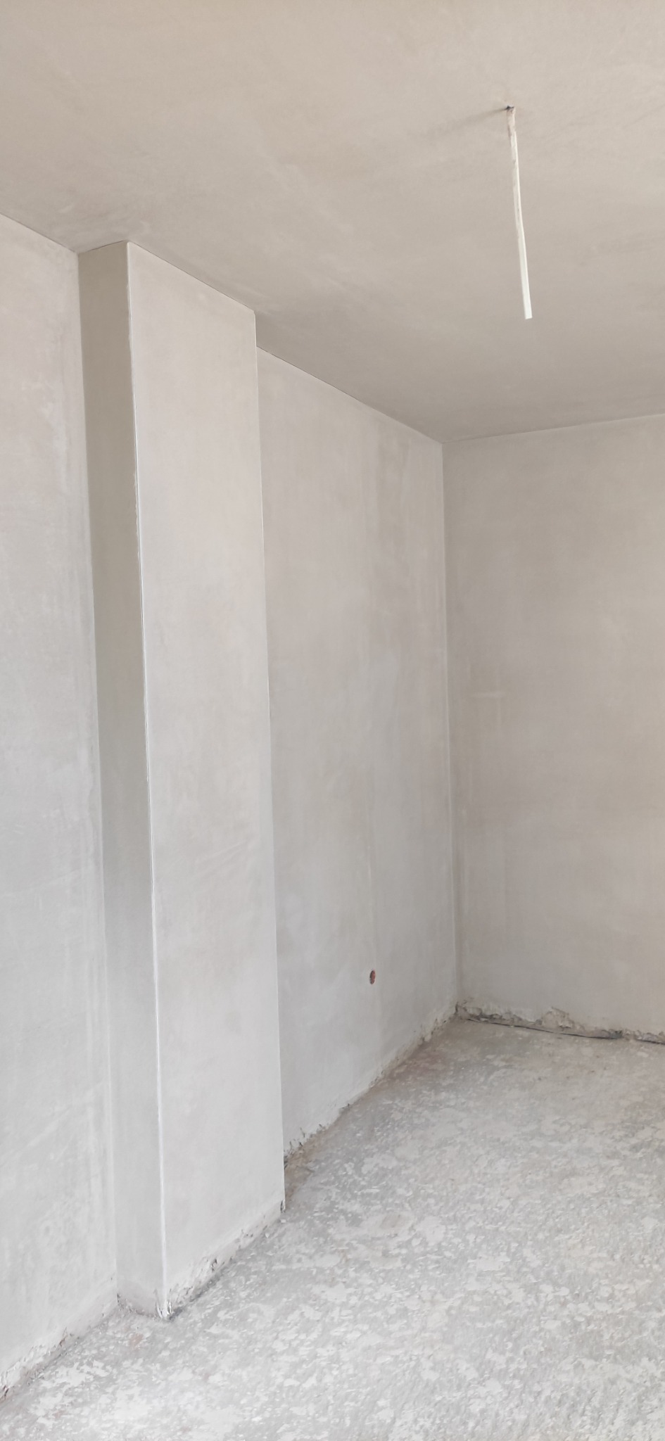 Surowe, jasne wnętrze po nałożeniu tynku, widoczny pionowy element konstrukcyjny oraz niedoskonałości na podłodze i ścianach.