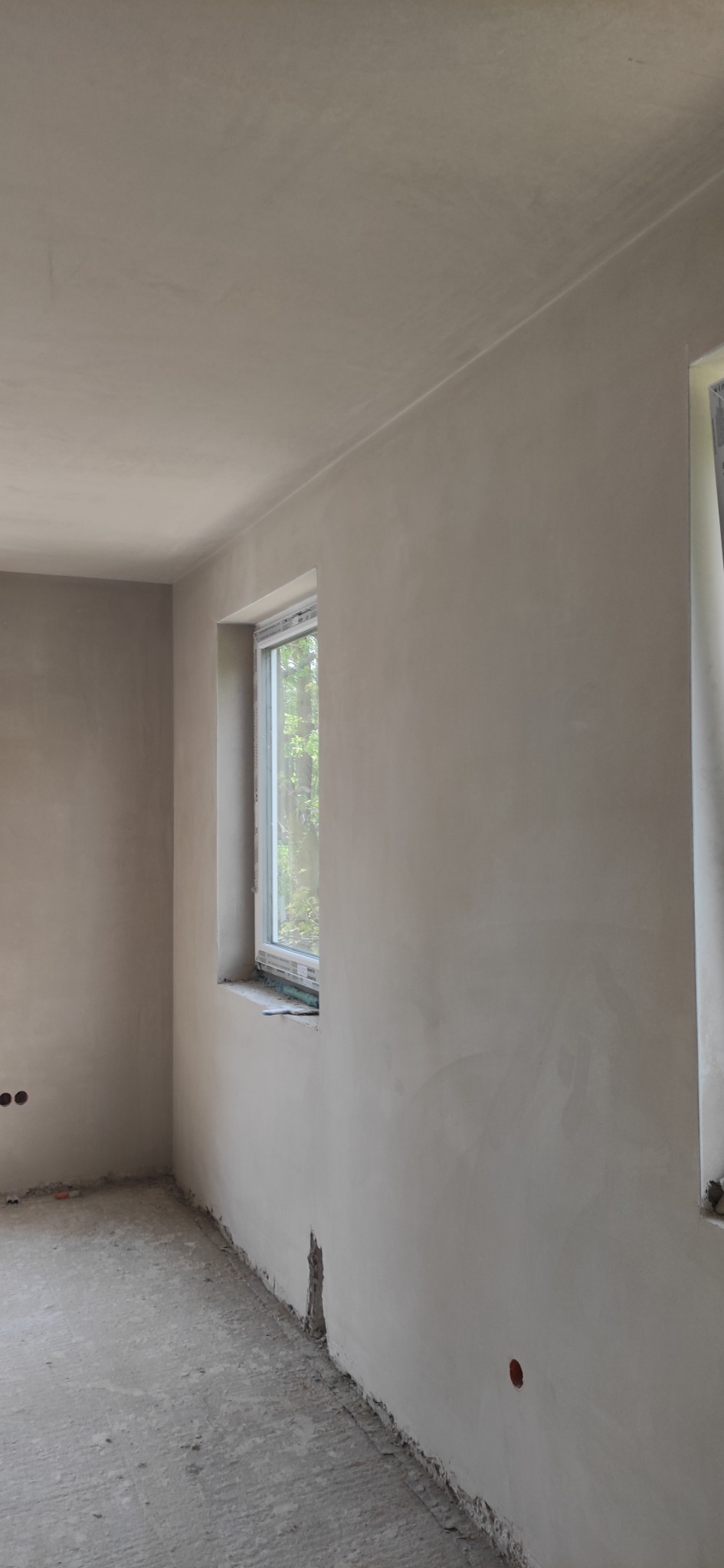 Wnętrze pomieszczenia po nałożeniu szarej warstwy wykończeniowej na ściany i sufit, widoczne okno z widokiem na zieleń, surowy beton na podłodze, ślady niedokończonych prac przy listwach...