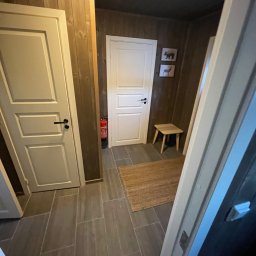 Jakub Parkiety-Malowanie - Wnętrze korytarza z drewnianymi ścianami i szarą podłogą z płytek, widoczne białe drzwi z czarnymi klamkami, mały stołek, dywanik i gaśnica.