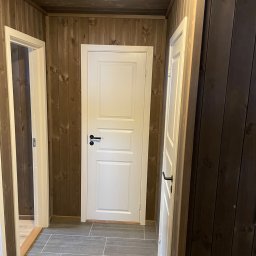 Jakub Parkiety-Malowanie - Korytarz wykończony drewnianą boazerią z dwiema białymi, zamkniętymi drzwiami z czarnymi klamkami oraz szarą podłogą z płytek imitujących kamień.