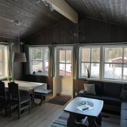 Jakub Parkiety-Malowanie - Wnętrze drewnianego domu z widokiem na zimowy krajobraz, drewniane panele na ścianach i suficie, stół z ławką, sofa i stolik kawowy z pilotem i świecą.